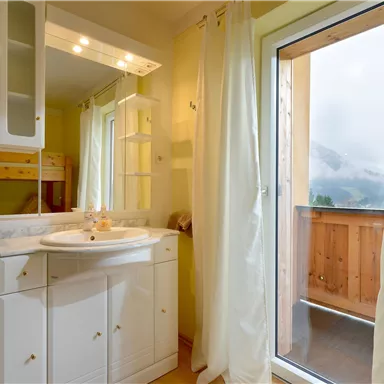 Ein helles Badezimmer mit einem Waschbecken und einem großen Fenster. Durch das Fenster ist ein Balkon mit Bergblick zu sehen.