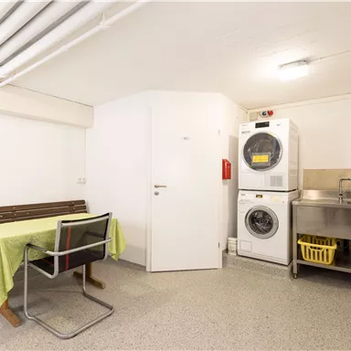 A simple laundry room with washing machines and a stainless steel sink. There is also a wooden bench and a table with a green tablecloth.
