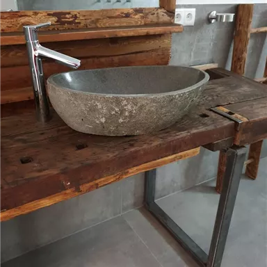 Ein moderner Waschtisch aus Holz mit einem eleganten Steinschalenbecken. Im Hintergrund sind graue Fliesen und rustikale Holzdetails zu sehen.
