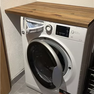 A modern washing machine with a wooden surface. It stands in a bright room on gray tiles.
