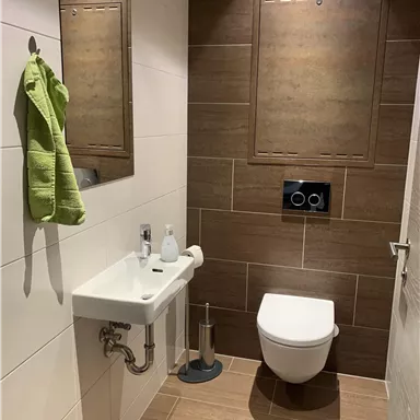 Eine moderne Toilette mit braunen Fliesen und einem kleinen Waschbecken. Ein grünes Handtuch hängt an der Wand.