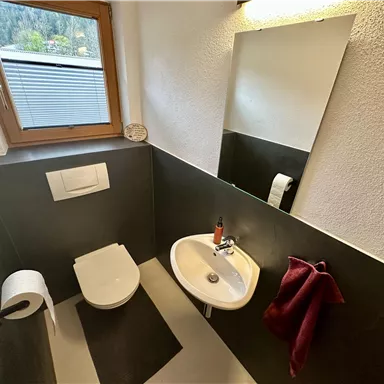 Ein modernes Badezimmer mit einer Toilette, einem Waschbecken und einem Spiegel. Die Wände sind in dunklen Tönen gehalten und es gibt ein Fenster, das Tageslicht hereinscheinen lässt.