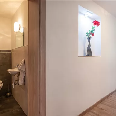 Ein heller Flur mit einem Badezimmer auf der linken Seite. An der Wand ist eine Vase mit einer roten Rose dekoriert.