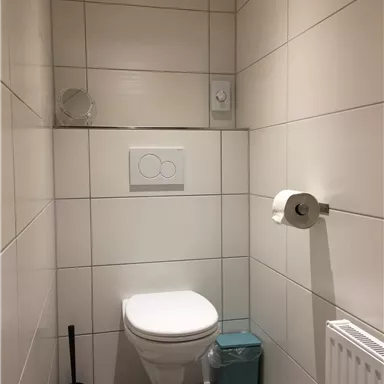 Ein modernes Badezimmer mit weißen Kacheln und einer Toilette. Neben der Toilette befindet sich ein Toilettenpapierhalter und ein kleiner Mülleimer.