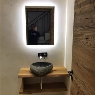 Ein modernes Badezimmer mit einem ovalen Waschbecken aus Stein und einem Holzregal. Der beleuchtete Spiegel und die Holzdetails verleihen dem Raum eine warme Atmosphäre.