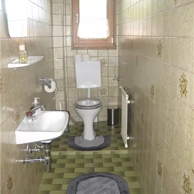 Ein kleines Badezimmer mit einer Toilette, einem Waschbecken und einem Fenster. Die Wände sind gefliest und der Boden hat ein grünes Muster.