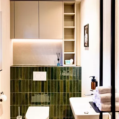 A modern bathroom with green tiles, an elegant toilet, and a sink. The walls are light, and there are practical shelves for storage.