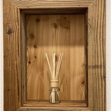 A decorative wooden frame with a light wood panel in the background. In the center stands a bottle with fragrance sticks.