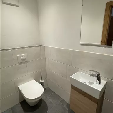 Ein modernes Badezimmer mit einer Toilette und einem kleinen Waschbecken. Die Wände sind hell gefliest und der Boden ist dunkel.