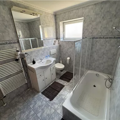 Ein modernes Badezimmer mit Dusche und Badewanne. Fliesen in Grau und ein großer Spiegel sorgen für eine helle Atmosphäre.