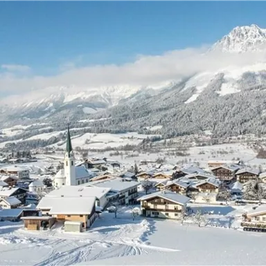 Eine verschneite Berglandschaft mit einem kleinen Dorf im Vordergrund. Der Himmel ist klar und blau, und die Umgebung ist von schneebedeckten Bergen umgeben.