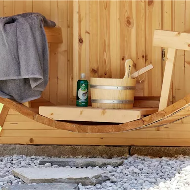 Ein gemütlicher Saunabereich mit Holzstruktur. Im Vordergrund befinden sich ein Handtuch, ein Eimer und eine Flasche Shampoo.