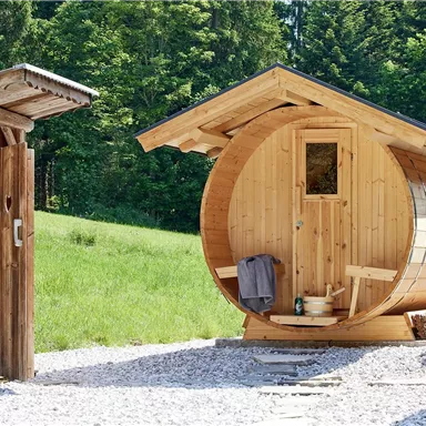 Eine moderne, runde Holzsauna steht neben einer kleinen Holzhütte. Im Hintergrund sind grüne Wiesen und Bäume zu sehen.