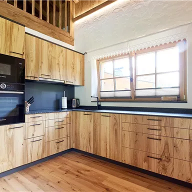 Eine moderne Küche mit Holzschränken und einer eleganten Arbeitsplatte. Große Fenster lassen viel Tageslicht herein.