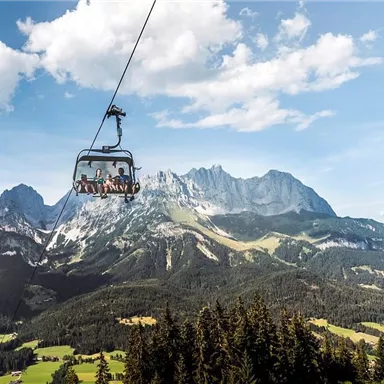 A cable car travels over green meadows and majestic mountains. The sky is blue with some clouds.