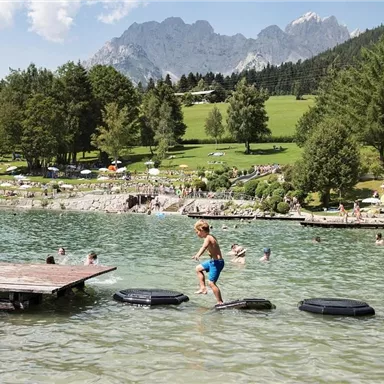 Ein Junge springt auf Bojen im klaren Wasser eines Sees. Im Hintergrund sind grüne Wiesen und Berge sowie viele Menschen beim Baden und Entspannen zu sehen.
