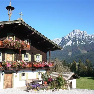A traditional wooden house with blooming flowers on the balconies. In the background, majestic mountains can be seen.