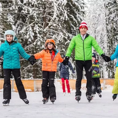 A cheerful group of children and an adult is enjoying ice skating on a frozen surface in the snow. They are all wearing colorful winter jackets and helmets.