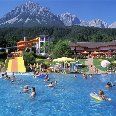 Ein Schwimmbad mit bunten Rutschen und vielen Menschen, die sich im Wasser vergnügen. Im Hintergrund sind beeindruckende Berge und eine sonnige Landschaft zu sehen.