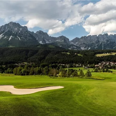 A green golf course landscape surrounded by majestic mountains. The sky is partly cloudy, and the place radiates tranquility.