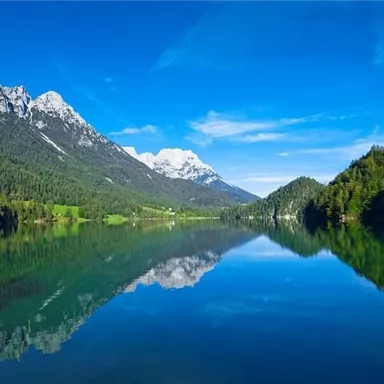 A clear lake surrounded by high mountains and lush greenery. The sky is blue and radiant, and the landscape is reflected in the water.
