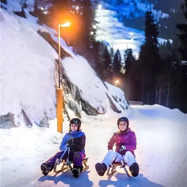 Two people are sitting on sleds, enjoying the nighttime winter landscape. The snowy road is illuminated by lanterns, while the mountains are visible in the background.