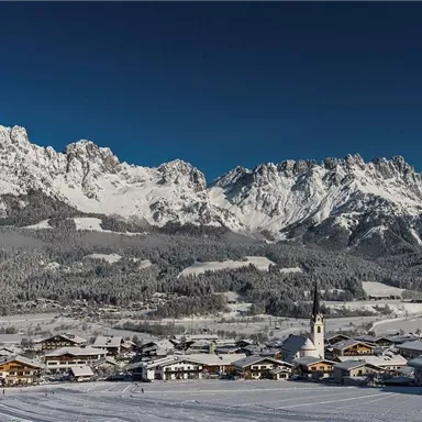 A picturesque village in a snow-covered landscape, surrounded by majestic mountains. The clear blue sky completes the winter scene.
