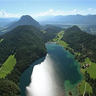 An impressive landscape with a sparkling lake, surrounded by green hills and mountains. The sky is clear and bright blue.