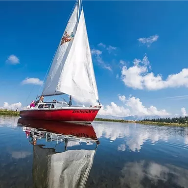 A sailboat with a white sail glides over a calm lake. The sky is blue with some clouds and reflects in the water.