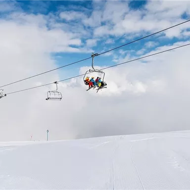 Eine Skiliftfahrt über eine verschneite Landschaft. Weiße Pisten und ein blauer Himmel mit Wolken sind zu sehen.