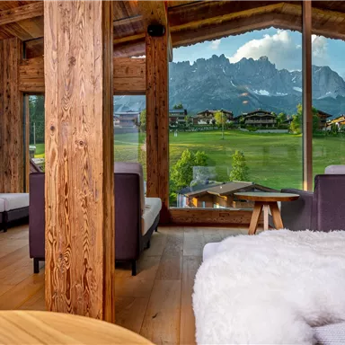Ein modernes, gemütliches Wohnzimmer mit großen Fenstern und Blick auf die Berge. Die Holzelemente und die sanfte Einrichtung schaffen eine einladende Atmosphäre.