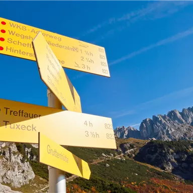 Ein Wegweiser mit mehreren Richtungen steht in einer bergigen Landschaft. Im Hintergrund sind beeindruckende Berge und ein blauer Himmel zu sehen.