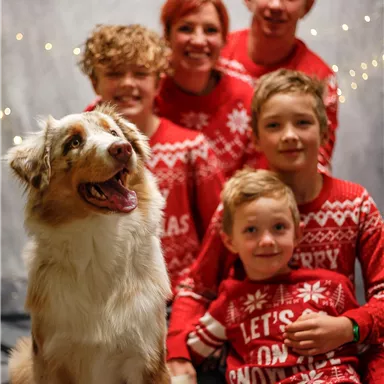 Eine fröhliche Familie posiert zusammen mit einem Hund vor einer festlichen Kulisse. Alle tragen weihnachtliche Pullover und lächeln.