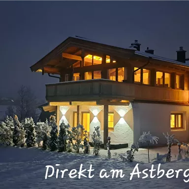 A cozy house in winter, lit by warm light. The snow and the surrounding trees create an inviting atmosphere.