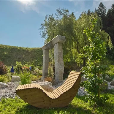 A beautiful garden with a modern wooden lounge chair and a stone sculpture. In the background, trees and colorful plants can be seen.