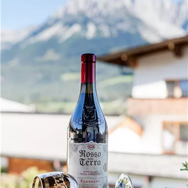 Eine Flasche Rotwein steht auf einem Tisch mit einer Weinglas daneben. Im Hintergrund sind die Berge und ein blauer Himmel zu sehen.