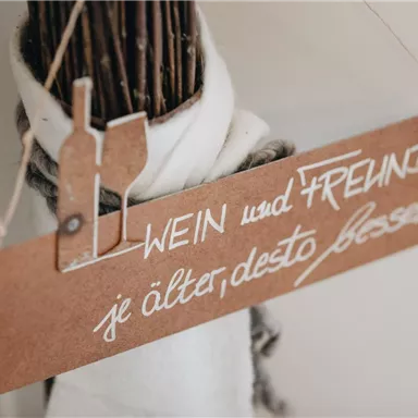 Ein dekoratives Holzschild mit der Aufschrift "WEIN und FREUNDE, je älter, desto besser." Es zeigt eine stilvolle und einladende Atmosphäre.