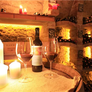 Ein gemütlicher Weinlagerraum mit Kerzenlicht. Auf einem Holzfass stehen zwei Weingläser und eine Flasche Rotwein.