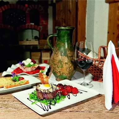 Ein schön angerichteter Tisch mit einem Gericht aus Fleisch, frischem Gemüse und einer Beilage. Daneben stehen eine Karaffe, ein Glas Rotwein und Servietten.