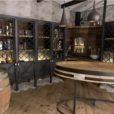 Ein gemütlicher Weinkeller mit dunklen Regalen und einem runden Holztisch. Im Hintergrund sind Weinfässer und dekorative Elemente zu sehen.