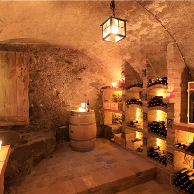 Ein gemütlicher Weinraum mit Holzregalen, die mit Flaschen gefüllt sind. Sanftes Licht von Kerzen und einer Lampe sorgt für eine einladende Atmosphäre.