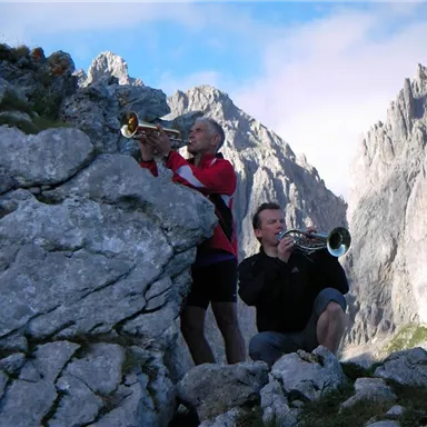Two men are playing trumpet on a rock in the mountains. In the background, impressive mountain peaks and a clear sky can be seen.
