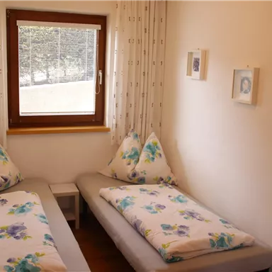 Ein helles Schlafzimmer mit zwei Einzelbetten. Die Betten sind mit blütenbedruckten Bettlaken und Kissen dekoriert, und ein Fenster bietet einen Blick nach draußen.