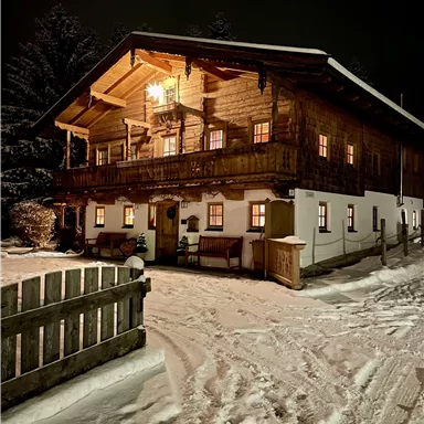 A cozy wooden house in the snow at night, illuminated by warm light. The path to the house is snow-covered and offers an inviting atmosphere.