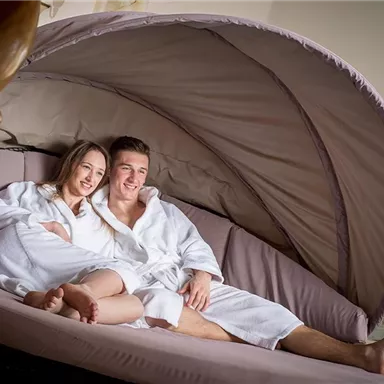 Ein Paar in Bademänteln entspannt auf einer bequemen Liege. Sie lächeln und genießen die gemütliche Atmosphäre.