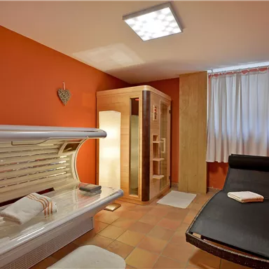 Ein gemütlicher Wellnessbereich mit einer Sonnenbank und einem Entspannungsliege. Die Wände sind in warmen Orangetönen gehalten und durch Fenster mit Vorhängen sanft beleuchtet.