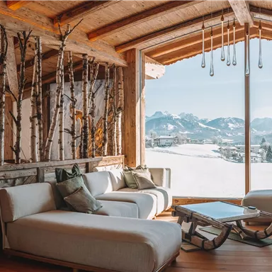 Eine moderne Innenausstattung mit bequemen Sofas und natürlichem Holz. Der Blick durch große Fenster zeigt eine malerische Berglandschaft und schneebedeckte Hügel.