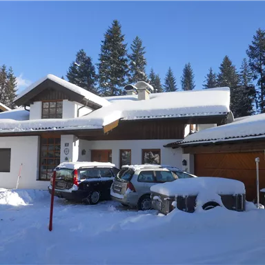 Ein modernes Haus in einer Schneelandschaft mit hohem Schnee. Im Hintergrund sind Nadelbäume und blauer Himmel zu sehen.