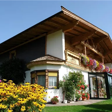 A charming house with a wooden balcony and colorful flowers. The garden is lush and green, surrounded by mountains in the background.