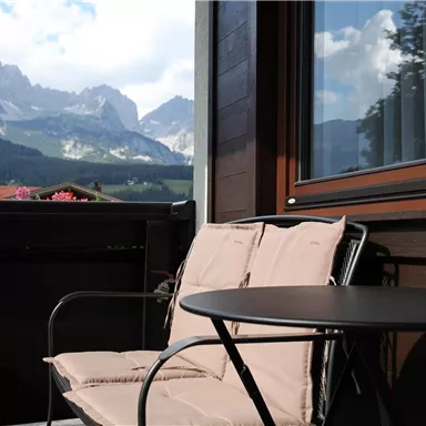 A cozy balcony with a chair and a small table. In the background, impressive mountains and a clear sky can be seen.